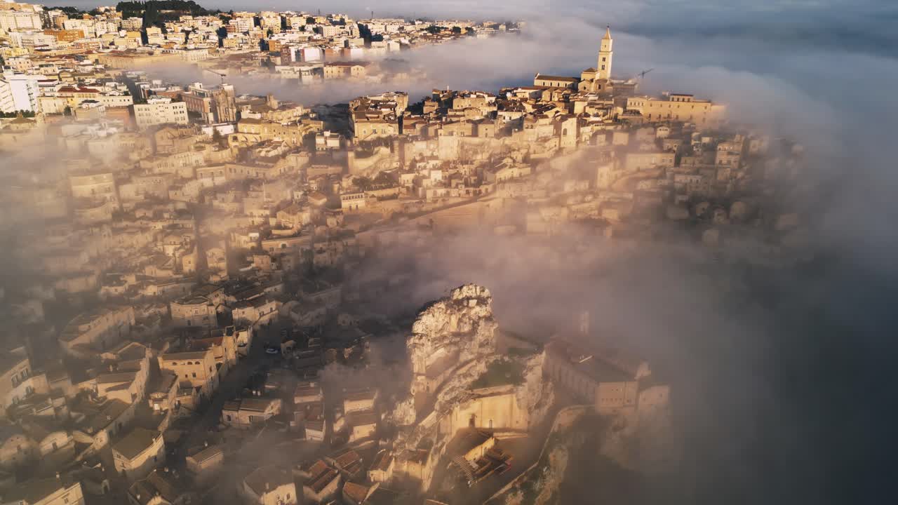 drone shot of old Italian city town - Matera. The city is shrouded with low handing clouds, buildings are visible through them. Warm colours of the rising sun reflected against buildings
