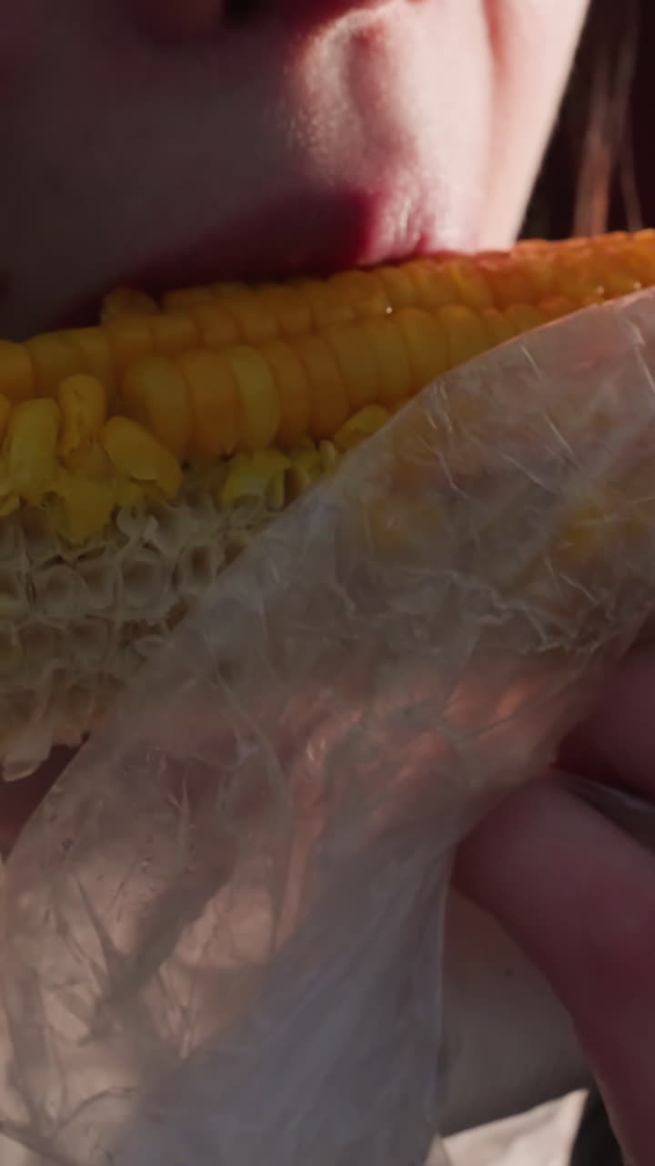 primer plano de una niña comiendo maíz sostenido por un palo envuelto en plástico blanco con luces bokeh en un fondo borroso, vapor que se eleva del maíz fresco en un entorno al aire libre, disfrutando de un bocadillo de comida callejera