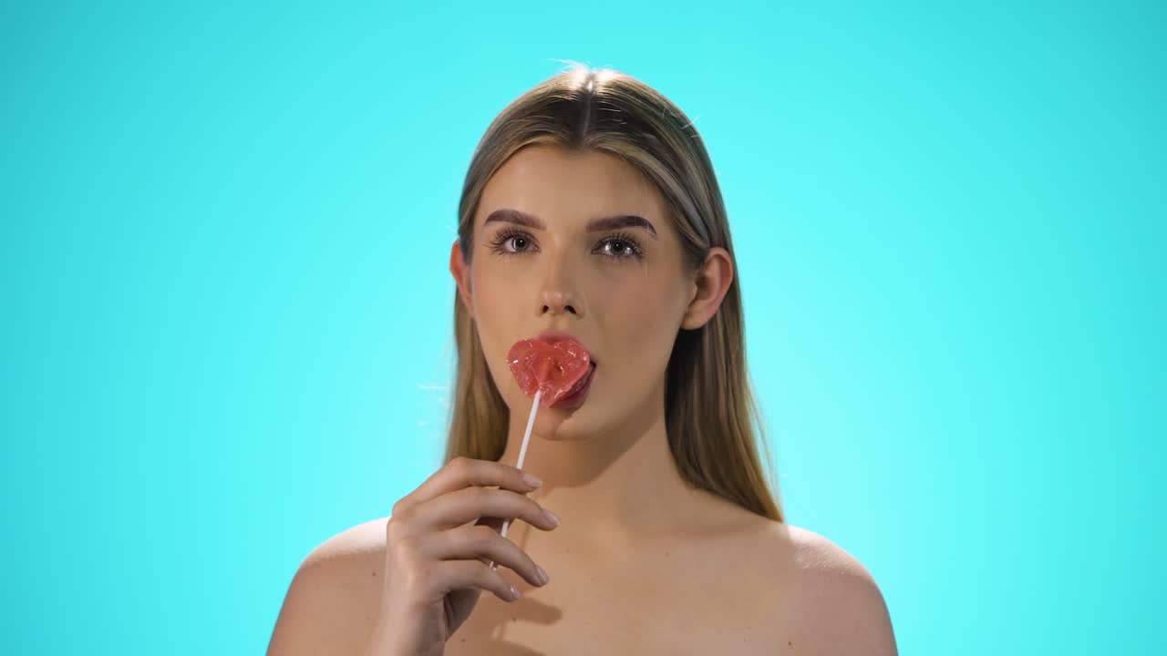 Medium Static slow motion shot of a young caucasian naked woman licking a heart shaped lollipop while looking sexy into the camera in slow motion against turquoise background