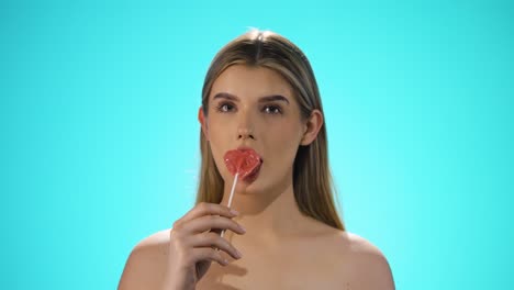 medium static slow motion shot of a young caucasian naked woman licking a heart shaped lollipop while looking sexy into the camera in slow motion against turquoise background