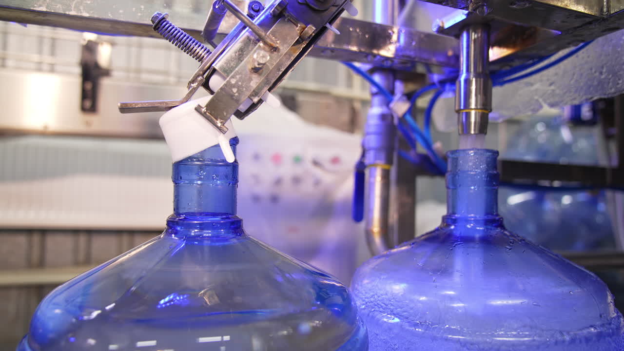 Water is poured into big blue bottle and overflows it. Filled bottles are covered with plastic white corks. Close up.