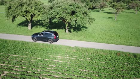 A-Volvo-xc-90-car-drives-on-a-small-bumpy-road-in-between-fields,-few-trees,-Swiss-countryside,-Vaud,-drone-aerial-view