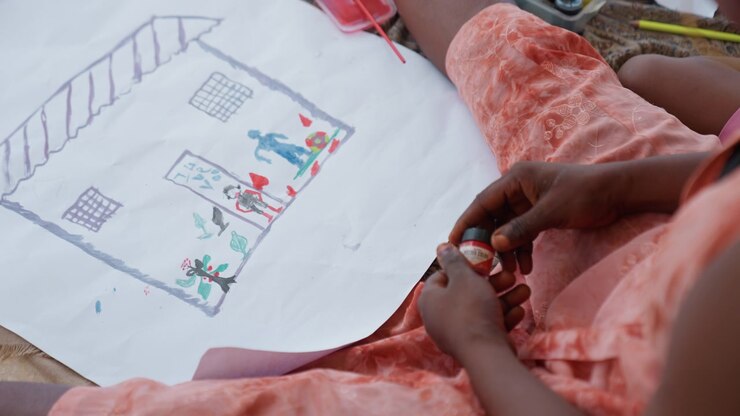 Child hands carefully apply red and black paint to details on colorful house drawing featuring people playing soccer gardening and interacting. scene focuses on precise movements of fingers
