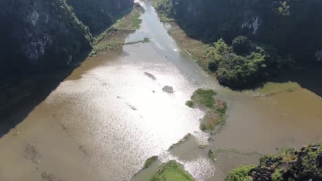 drone aerial view in vietnam vertical panning showing rocky mountains covered with green trees over a wide brown river in ninh binh on a sunny day