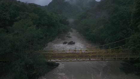 Man-in-yellow-coat-walks-over-bridge-at-Cahabon-river-Guatemala,-aerial