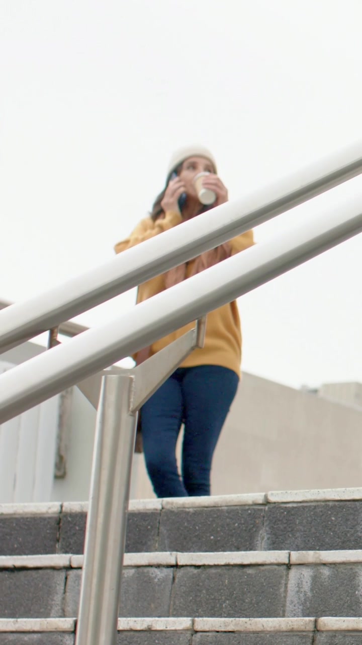 Video verticale: Una donna con un berretto risponde a una chiamata, scende le scale, tiene il telefono e sorseggia caffè.