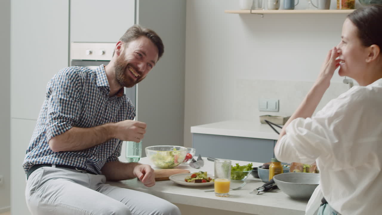 Laughing Couple Playing And Having Fun Together Sitting On Stool In A Modern Kitchen