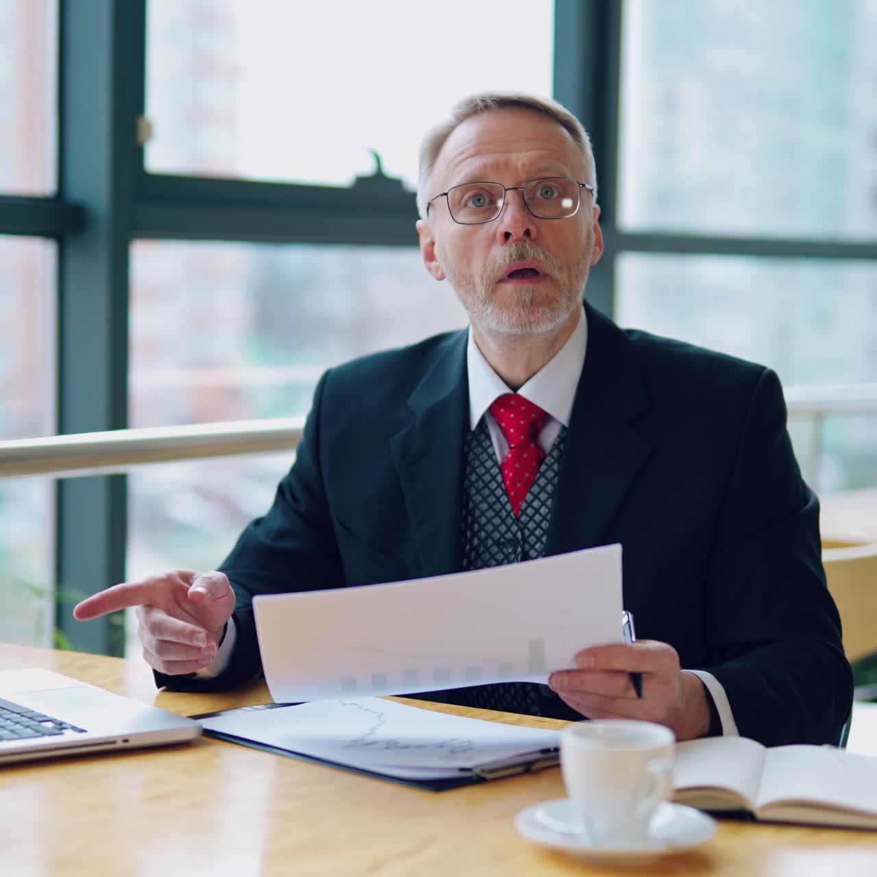 Businessman is checking a printed report or document while sitting at table, working on paper work at a laptop explaining his point of view. Video in an office.