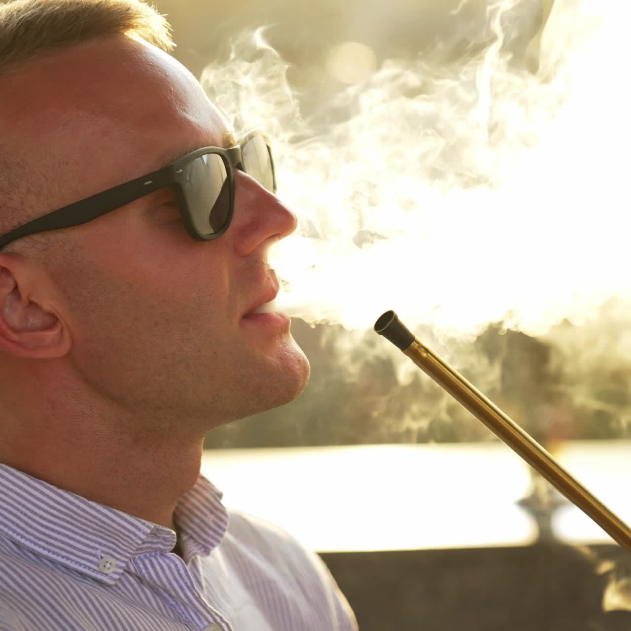 Blond man wearing sunglasses smoking hookah. Man produces smoke from nose and mouth at once. Close up