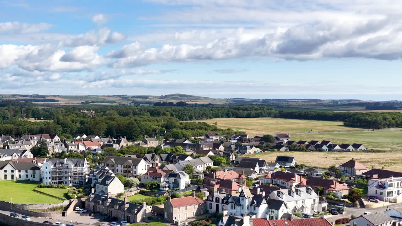 Drone pans above residential houses, countryside, and shoreline under bright daylight in Dundee, Scotland