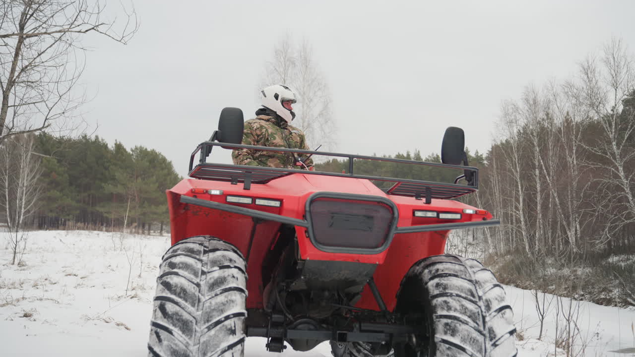 ATV rosso che sfreccia attraverso un campo innevato, pneumatici larghi che creano nuove tracce. Pilota con casco che accelera attraverso una radura delimitata da betulle. Cielo grigio e freddo, primo piano della griglia e delle sospensioni. Attività outdoor ad alta energia.