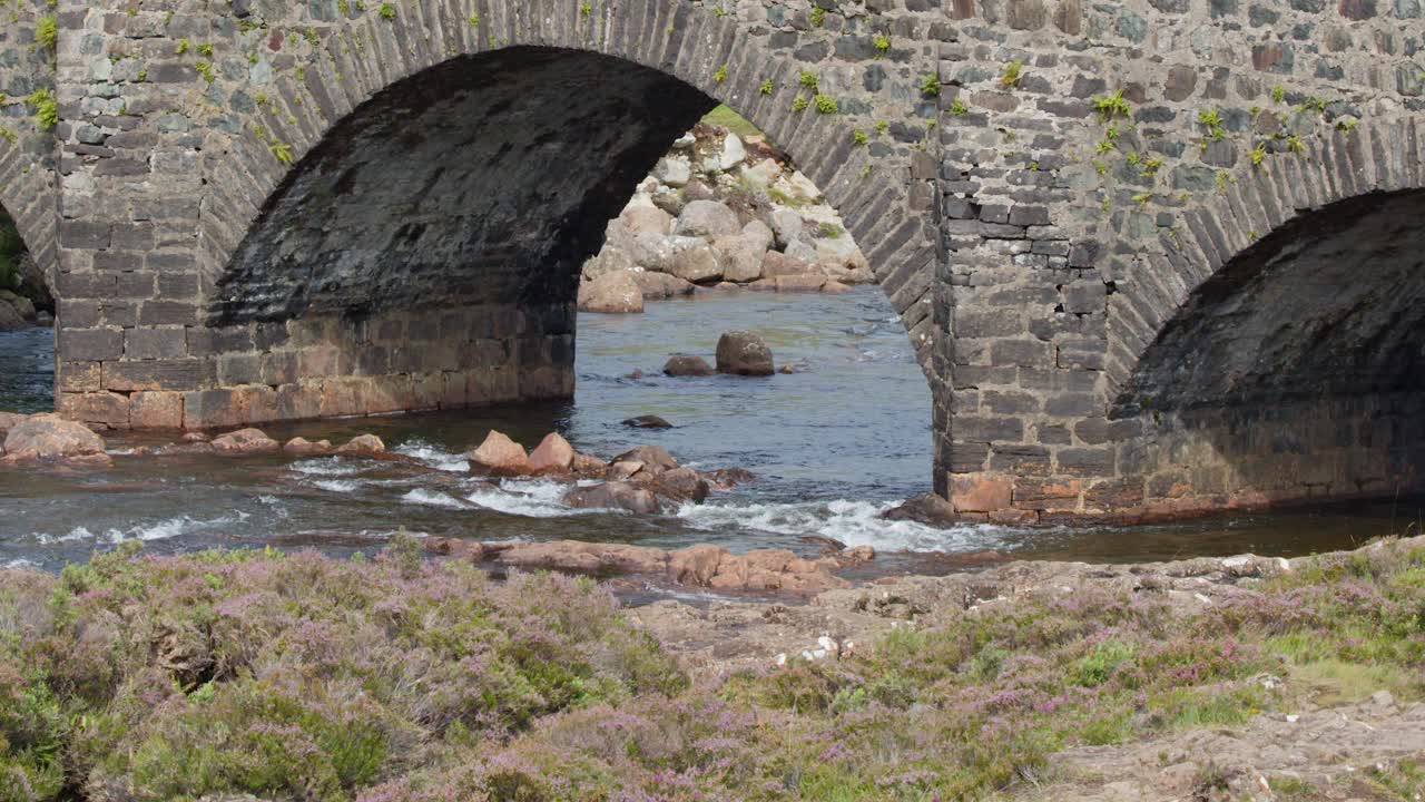 Tourists walk across old stone arch bridge above river, daylight, scenic rural Scottish Highlands landscape