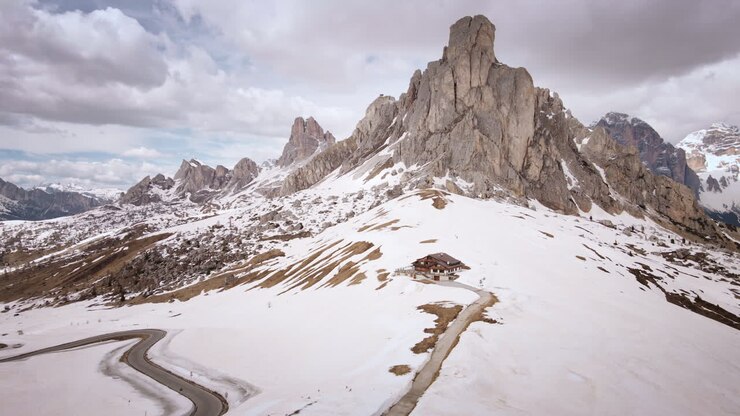 Aerial drone moves left over snowy Passo Giau road, rifugio, and rugged Dolomites peaks with wide panoramic mountain views in soft spring light