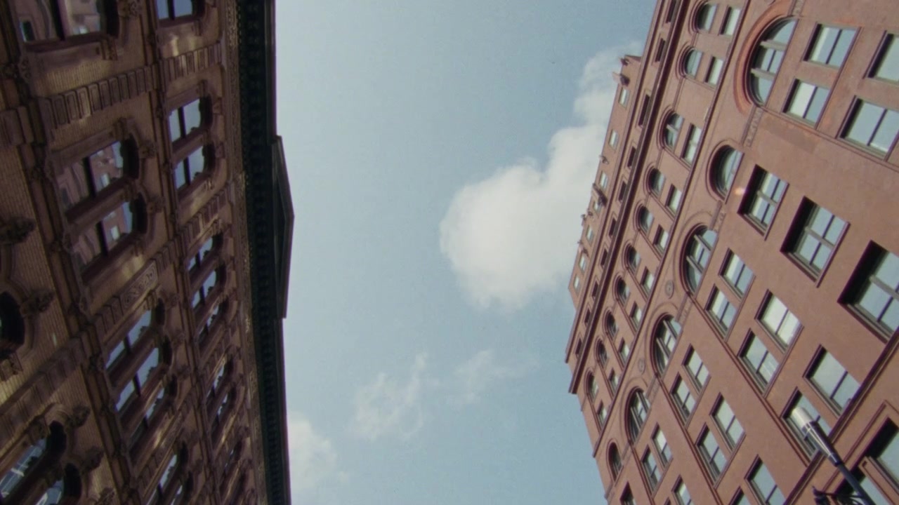 Vista stradale dal basso che si muove tra due edifici storici contro un cielo blu con nuvole, su pellicola 16mm.