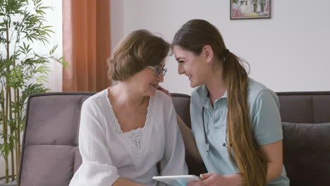 mulher sênior e médica sentada em um sofá conversando e rindo enquanto olha para um tablet 1