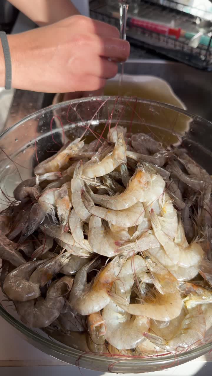 Closeup of woman’s hands washing fresh raw shrimp in bowl under running kitchen tap water