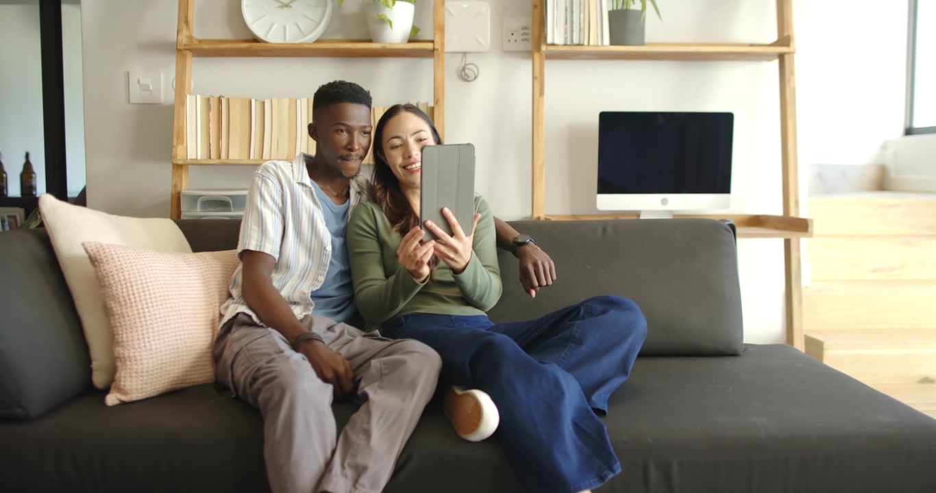 Couple sitting on grey sofa at home, woman tapping tablet, man leaning in reacting to content