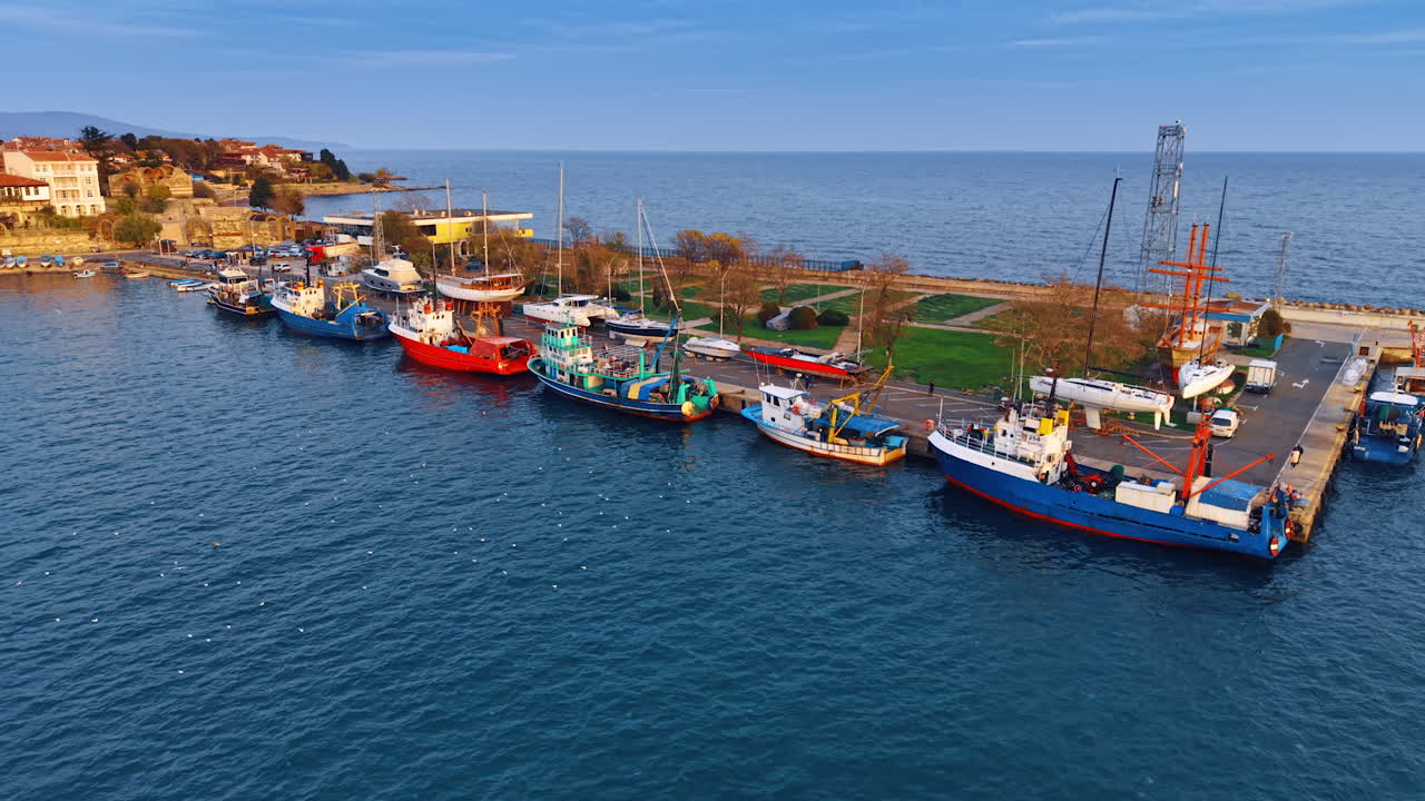 Diverse barche da pesca e cargo sono ormeggiate a un molo in un porto. L'ambientazione è nei pressi di una città costiera con edifici sullo sfondo.