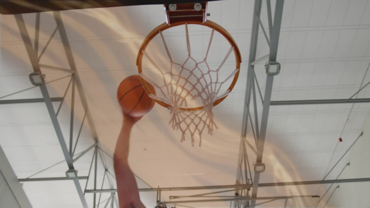 Braccio di un giocatore che afferra il pallone da basket e segna un canestro, mentre la rete si tende sotto il soffitto della palestra