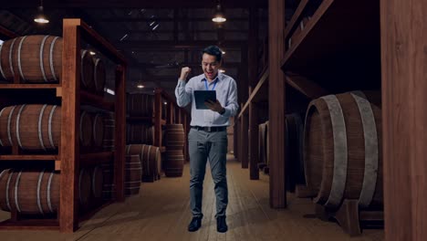 full body of an asian male professional worker standing with his tablet in cellar of winery, he raises his fist up with screaming goal after typed on the tablet