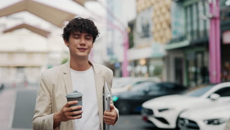 man with curly hair walking on street with coffee cup and laptop