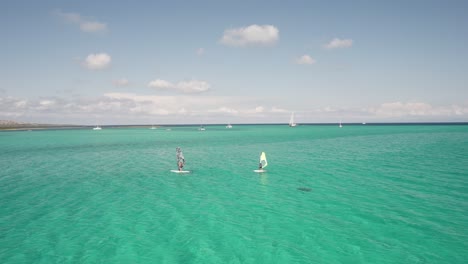 two windsurfers glide across clear turquoise waters under a sunny sky, aerial shot