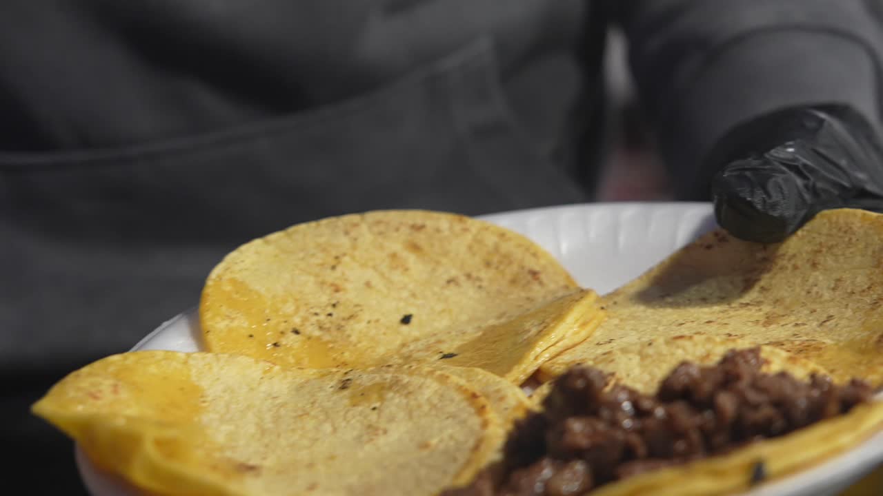 Primo piano di una persona che riempie dei tacos con carne su un piatto di carta, usando un cucchiaio. Il movimento rallentato evidenzia la consistenza, la freschezza e i vivaci sapori messicani del ripieno.