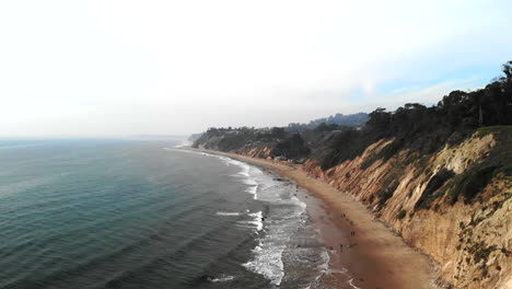 Drone-Aéreo-Que-Se-Eleva-Sobre-Las-Olas-Azules-Del-Océano-Pacífico-Frente-A-Los-Acantilados-De-La-Playa-En-La-Costa-Nublada-De-Santa-Bárbara,-California