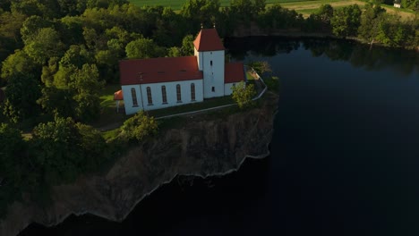 Smooth lateral drone flyby of the Wehrkirche Beucha, perched on a rocky cliff above a quarry lake in Saxony. Captured in soft sunset light, showcasing the dramatic edge and surrounding forest.