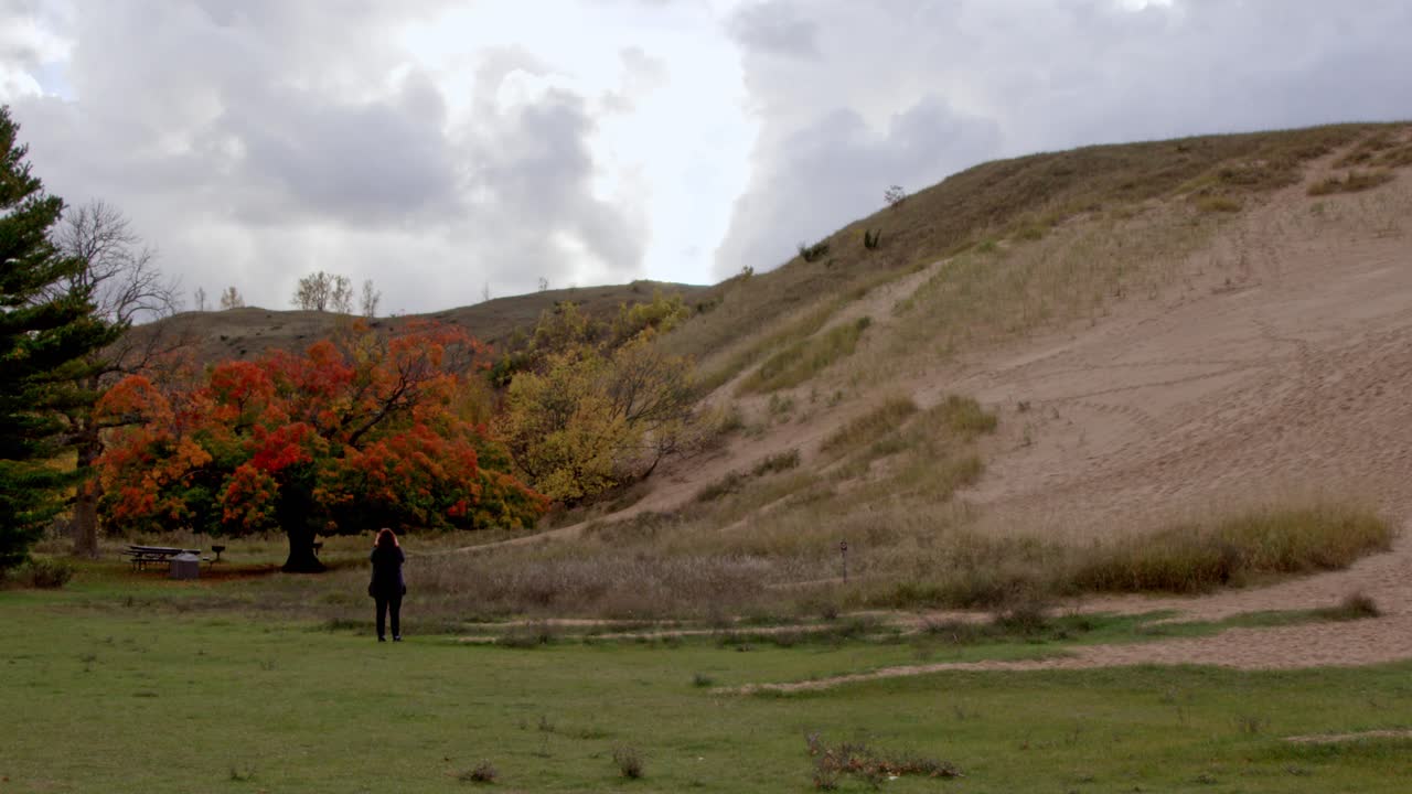Orta yaşlı bir kadın, bulutlu bir sonbahar gününde Michigan'daki Sleeping Bear Dunes Ulusal Göl Kıyısı'nda bulunan Dune Climb'in stabil video ile fotoğraflarını çekiyor