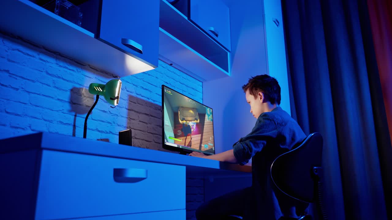 Boy playing video game at home. Concentrated gamer sitting at the desk in front of the computer and playing online games. Blue light in the room.