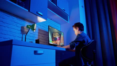 boy playing video game at home. concentrated gamer sitting at the desk in front of the computer and playing online games. blue light in the room.