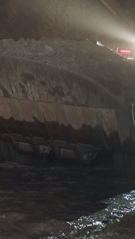 heavy mining machinery moves on dirty path washed with cold water. vehicle transports heap of ore in dipper illuminating way with bright light closeup
