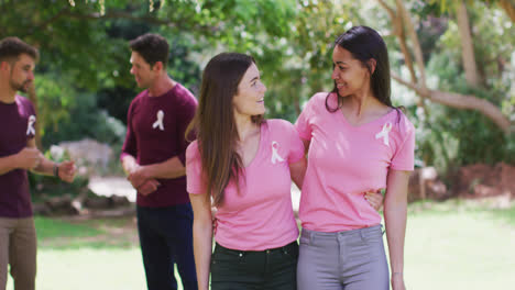 dos mujeres felices y diversas en camisetas rosas y cintas de cáncer, abrazando y flexionando los bíceps en el parque