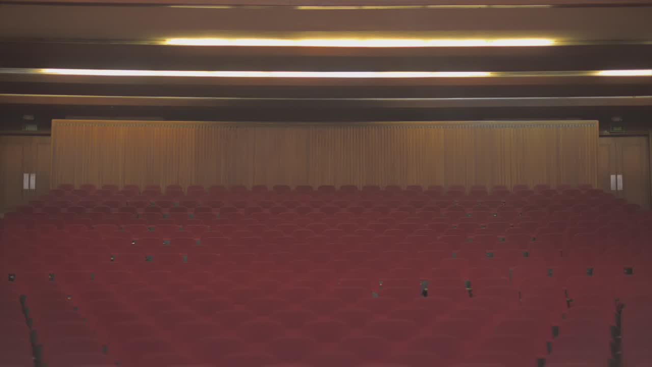 Tilt down of an abandoned theatre. The lights are on, and when the cameras gets to the end of the scene it reveals a lot of empty red chairs