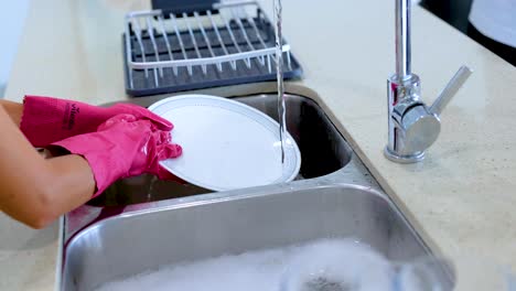 hands in pink gloves wash dishes under running water in a stainless steel sink, showcasing a clean, organized kitchen environment