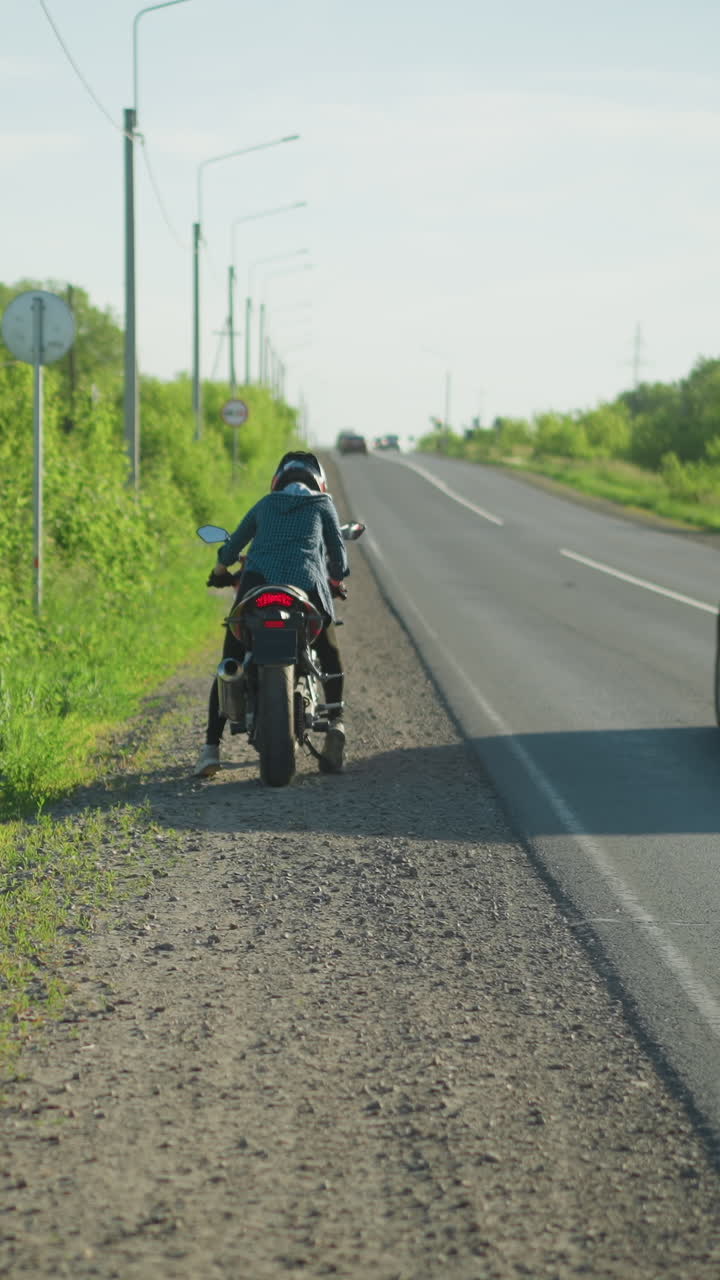 una vista trasera de una mujer subiendo a una motocicleta estacionada al lado de la carretera, con arbustos verdes exuberantes en el fondo y coches acercándose por detrás a lo largo de la carretera