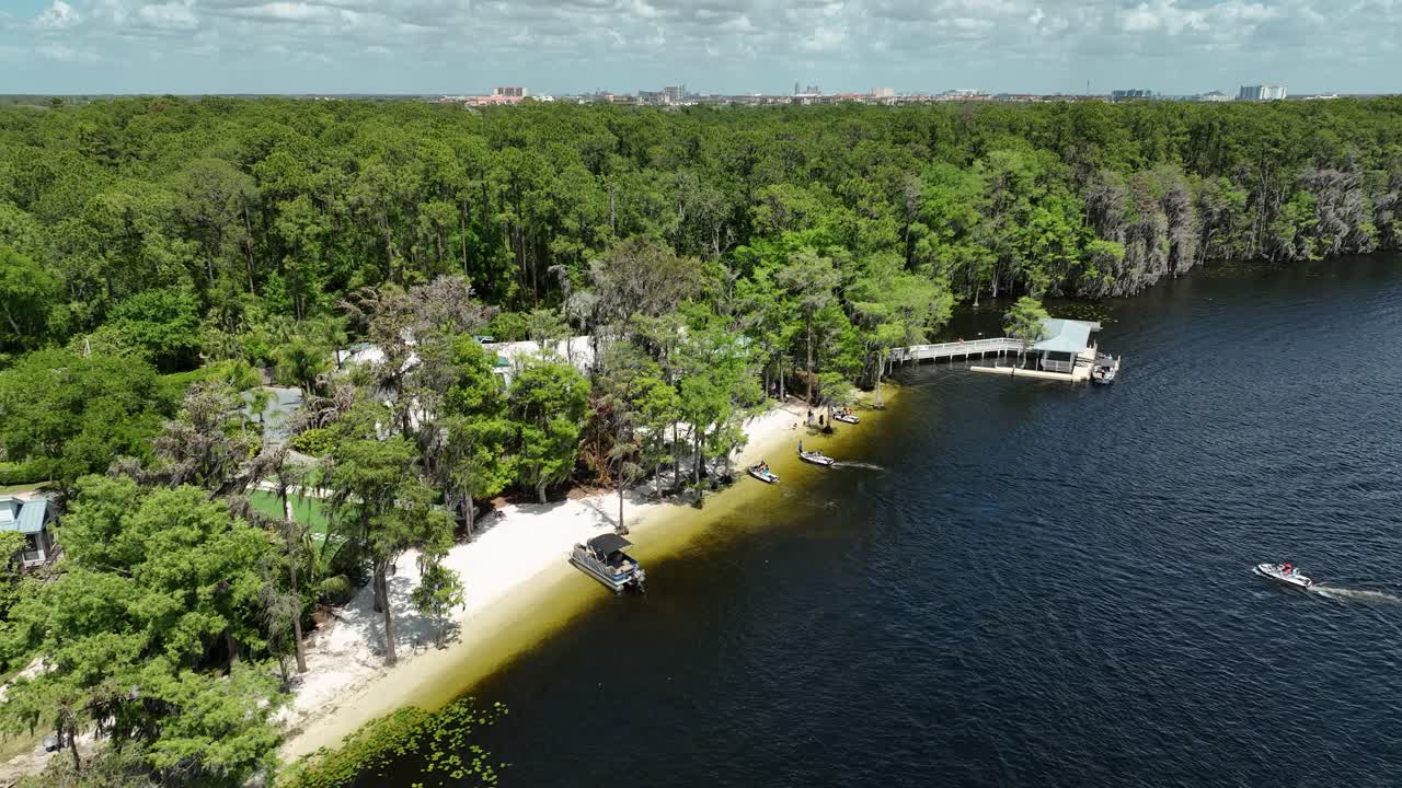 Tomada de drone ensolarada de Paradise Cove no Lago Bryan em Orlando, Flórida — barcos deslizando sobre águas calmas, florestas verdes exuberantes e um charmoso refúgio à beira-mar aninhado entre as árvores