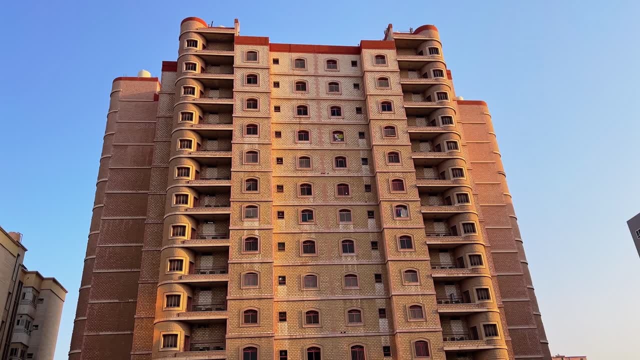 Vista frontal de um alto edifício residencial de apartamentos com fachada de tijolo, múltiplas varandas e janelas simétricas iluminadas pela luz solar quente contra um céu azul claro.