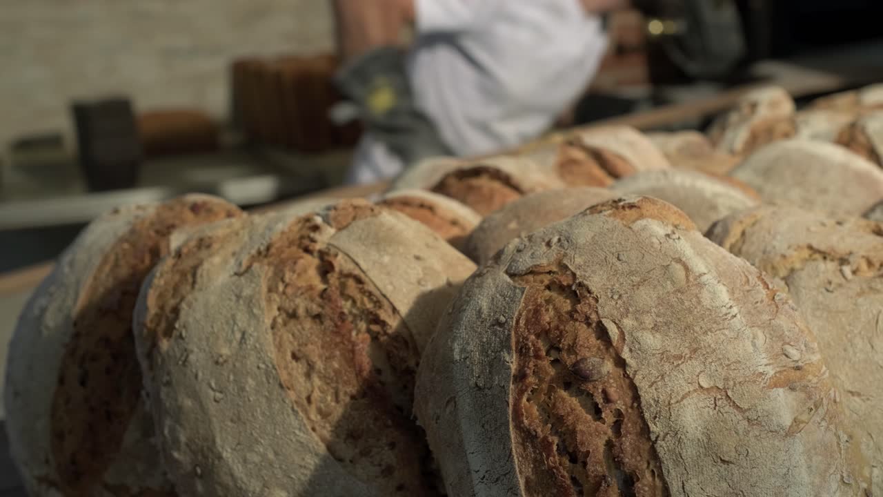 forground of pile of loaf bread, background of baker, with a baker’speel taking out more product