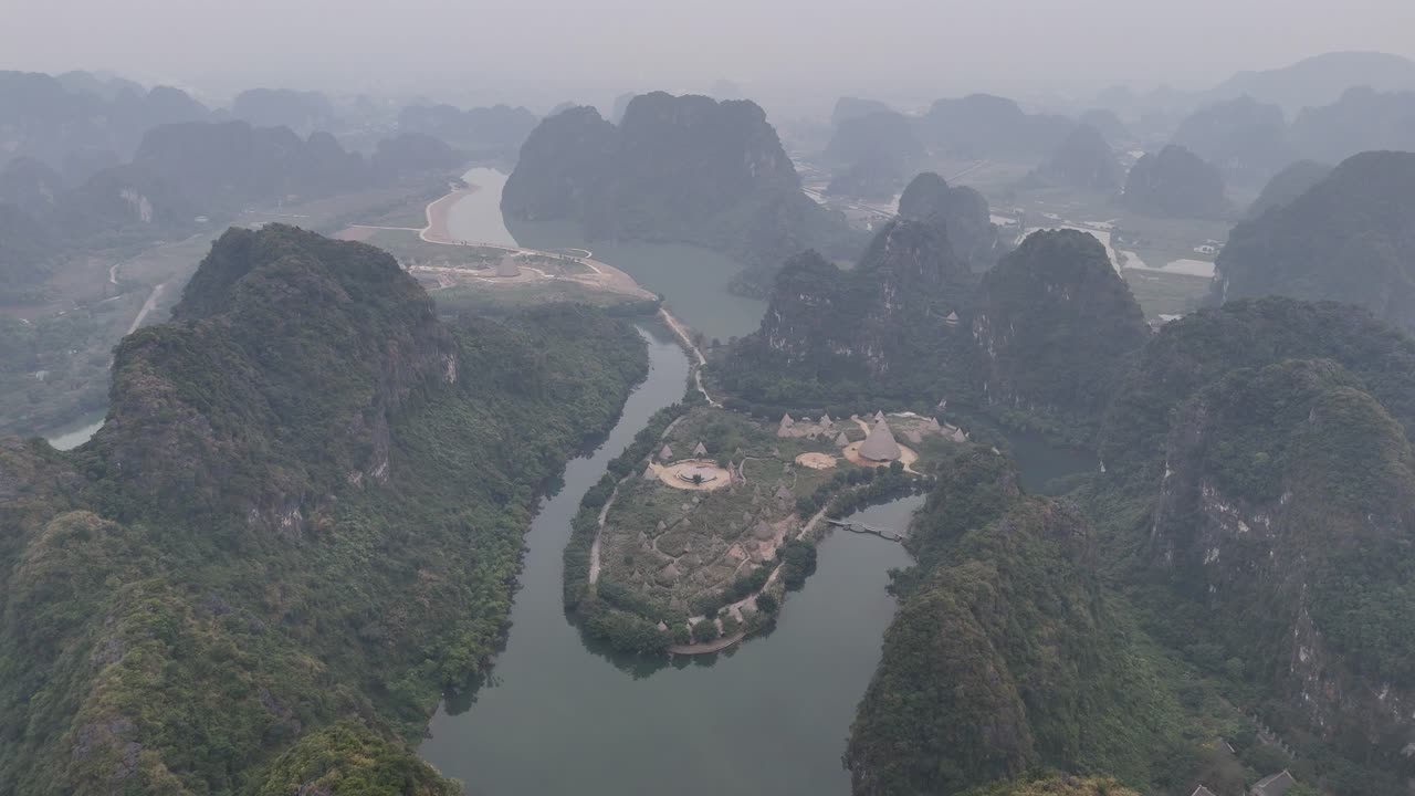 Drone video capturing the mist-covered limestone karsts and winding rivers of Vietnam. Lush greenery, dramatic terrain, and overcast sky create a serene, mystical atmosphere