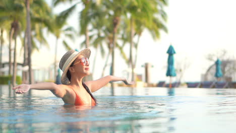 Stylish-Happy-Asian-Woman-With-Hat-and-Sunglasses-Raising-Hands-in-Swimming-Pool-on-Sunny-Summer-Day,-Slow-Motion