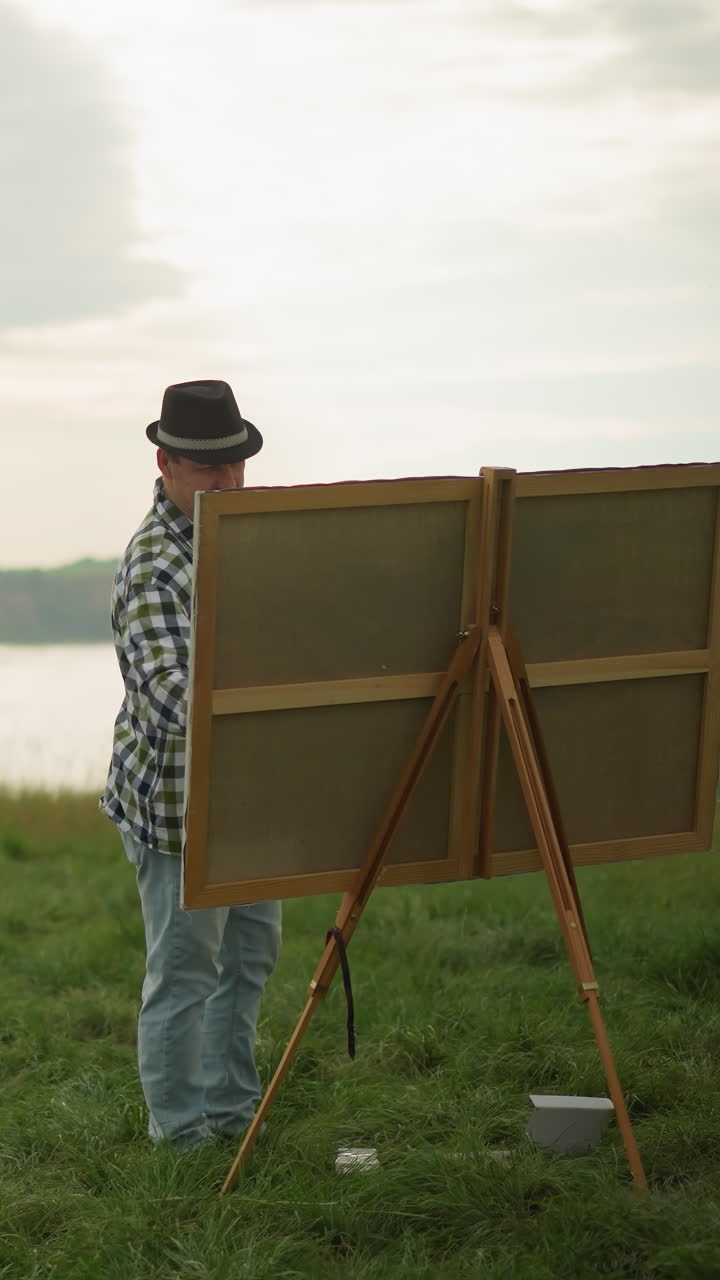 A sketcher in a black hat, checkered shirt, and jeans is engrossed in painting on a large canvas set up in a peaceful grass field by a serene lake. The calm setting, surrounded by natural beauty