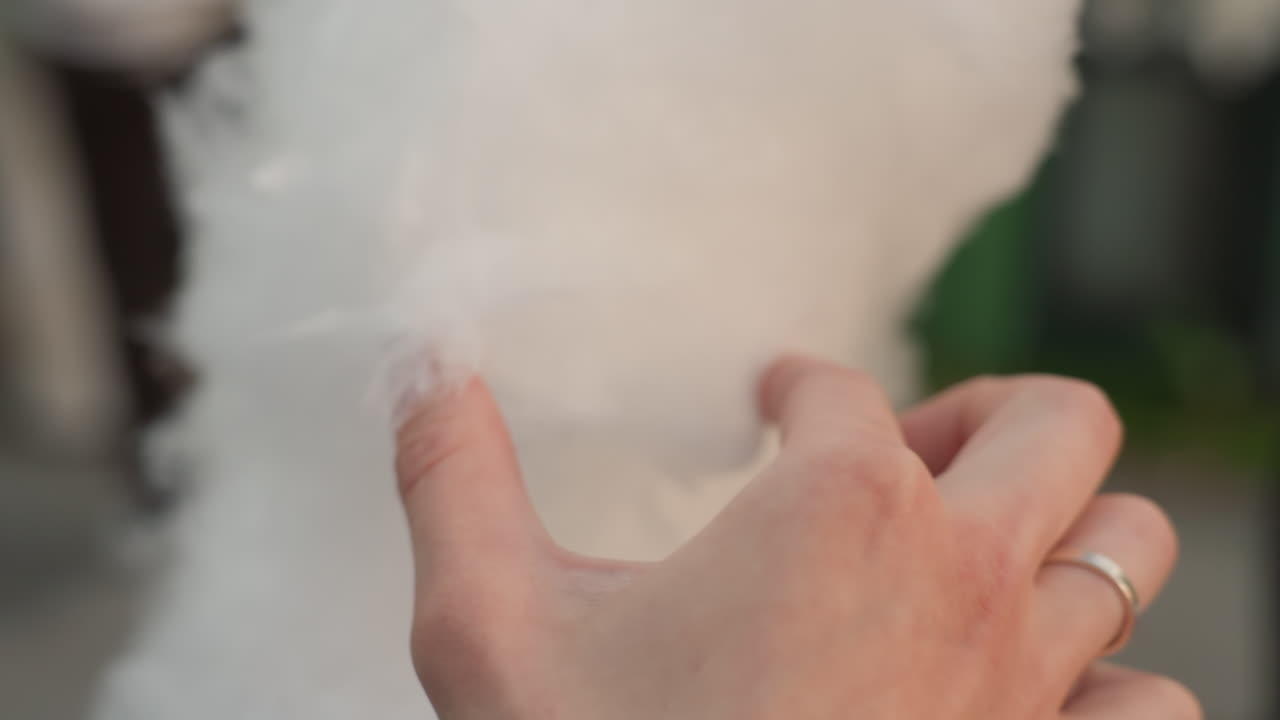 Nahaufnahme Zuckerwatte am Stiel, verheiratete Hand mit Ring zieht flauschige weiße Fäden auf einem Jahrmarkt im Freien, nostalgische Familienatmosphäre, Verkäufer im unscharfen Hintergrund, weiche luftige Textur, spielerischer Teilmoment
