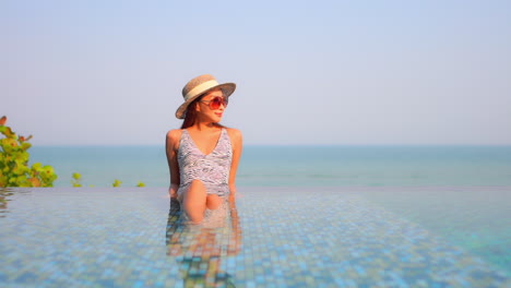 Mujer-Joven-Disfrutando-Del-Descanso-En-La-Piscina-Infinita-Al-Aire-Libre-Al-Atardecer-Usando-Sombrero-Para-El-Sol,-Gafas-De-Sol-Rojas-Y-Traje-De-Baño-Estilo-Monokini-Manchado