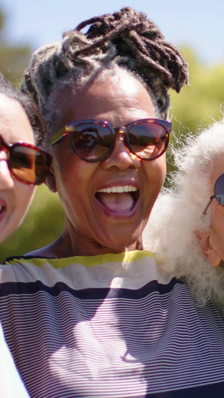 Vidéo verticale : Femme aux dreadlocks enlacée dans un parc, portant un haut rayé, ses lunettes de soleil reflétant un téléphone pour un selfie.