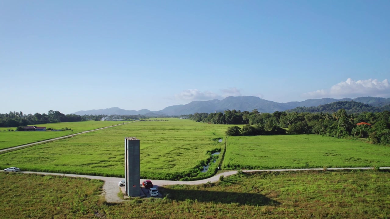 上昇する空撮視点から、マレーシアの田園地帯における熱帯稲作の壮大な規模が明らかになる。緑の水田が遠くの森林に覆われた山々へと広がる