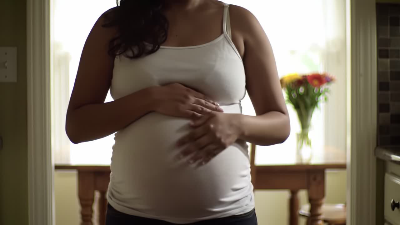 A pregnant woman stands in a warm kitchen, gently cradling her baby bump. Sunlight filters through the window, illuminating the colorful flowers nearby.