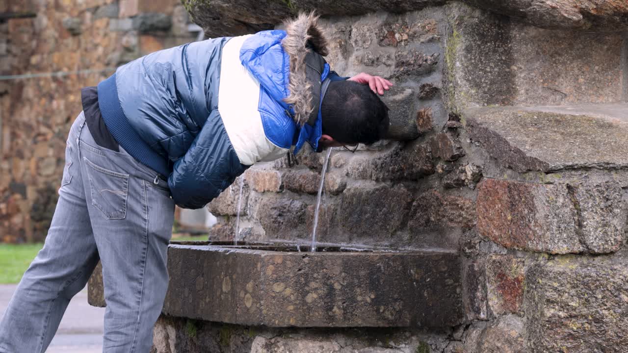joven con chaqueta ancha bebe del chorro de agua de una fuente de piedra en un pequeño pueblo rural