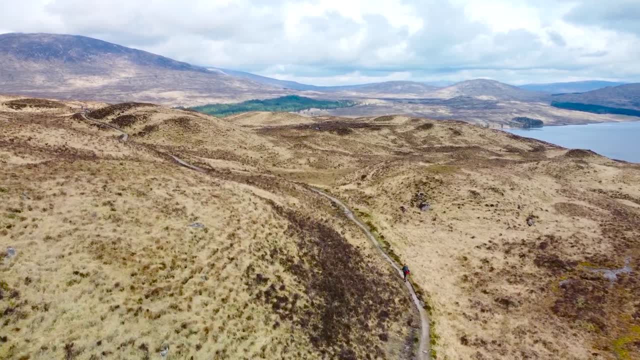 Aerial drone footage following and flying behind a hiker person walking on a rural road in scottish highlands landscape during a cloudy day. Large blue lake and mountains also visible in the back.